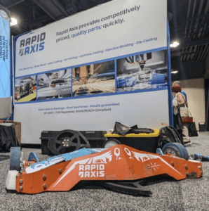 At A Trade Show Booth Boasting The Text &Quot;Rapid Axis Provides Competitively Priced, Quality Parts; Quickly,&Quot; An Orange Mechanical Part Reminiscent Of Battlebots Captures Attention, With The Company's Logo And Other Small Mechanical Objects Completing The Display.